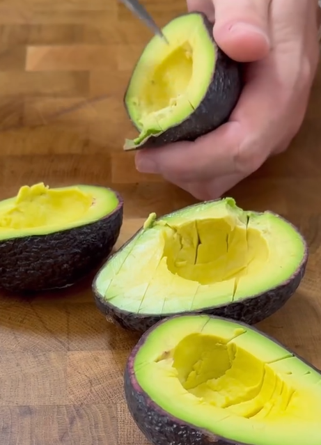 A hand holds an avocado half while scoring its flesh with a knife. Two scored avocado halves and one empty peel rest on a wooden cutting board.