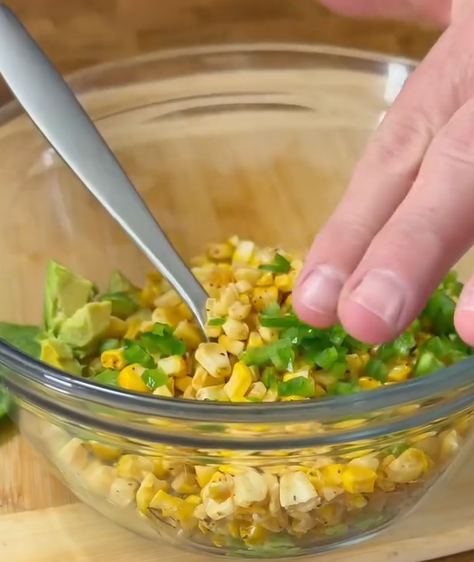 A hand sprinkling chopped green herbs over a glass bowl filled with a mixture of grilled corn, avocado, and seasonings; a spoon rests in the bowl.
