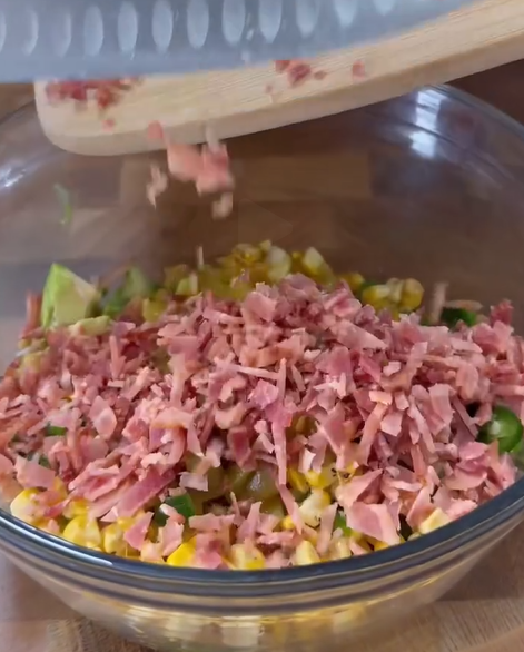 Chopped bacon is being added from a wooden cutting board into a glass bowl filled with corn, avocado, and other diced vegetables.
