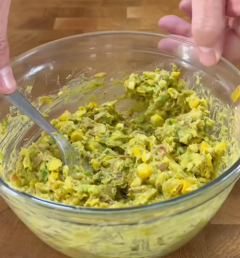A persons hands hold a glass bowl filled with a chunky avocado and corn mixture, being mixed with a spoon on a wooden surface.