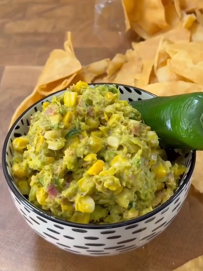 A bowl of guacamole mixed with corn sits on a wooden surface, topped with a jalapeño. Tortilla chips are scattered in the background.