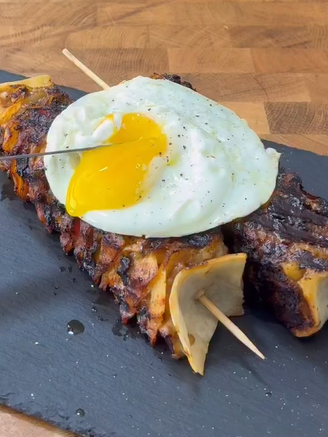 A grilled meat skewer with a fried egg on top, the yolk partially broken, served on a black slate plate over a wooden surface.