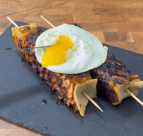 Two skewers of grilled meat with layered edges are topped with a sunny-side-up egg. The skewers are served on a black slate platter, and a knife is cutting into the runny egg yolk.