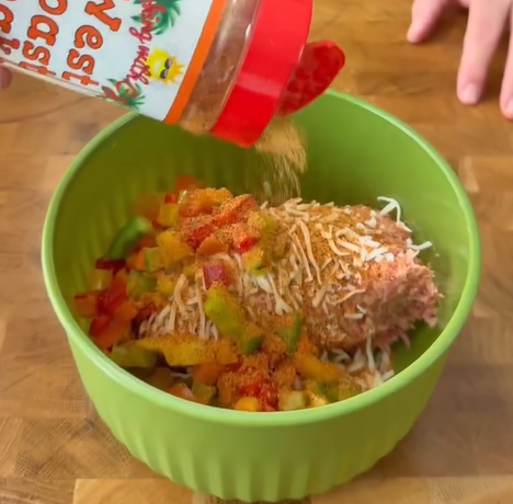 A hand sprinkles seasoning from a plastic jar onto a mixture of ground meat, shredded cheese, and chopped bell peppers in a green bowl on a wooden surface.
