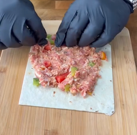 A person wearing black gloves is folding a sheet of dough filled with minced meat and chopped vegetables on a wooden cutting board.
