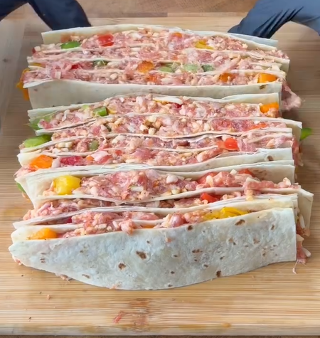 Several folded tortillas filled with a mixture of chopped meats, cheese, and colorful vegetables are lined up on a wooden cutting board, ready to be cooked or served.