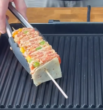 A hand uses tongs to place a skewer loaded with alternating pieces of marinated meat, vegetables, and flatbread onto a grill pan.