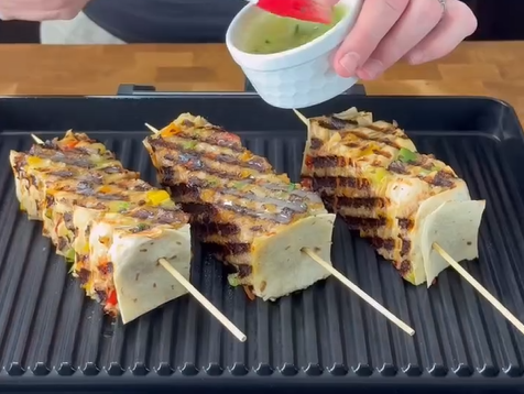 Three skewers with grilled paneer and vegetables are on a grill pan. A hand is pouring green sauce from a small white bowl over the skewers. The paneer pieces are charred with visible grill marks.