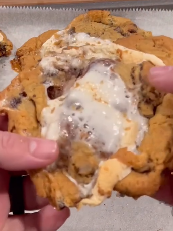 A close-up of hands breaking apart a large, gooey chocolate chip cookie with melted marshmallow filling, on a parchment-lined baking tray.