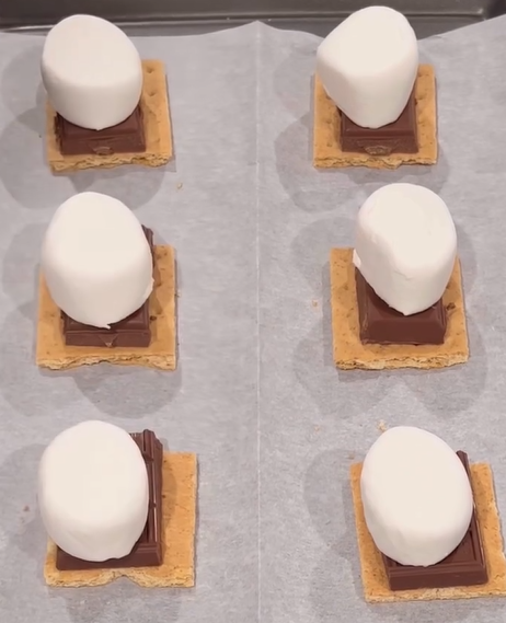 Six smores are arranged on a baking sheet. Each has a square graham cracker base, a piece of chocolate, and a large marshmallow on top, all placed on parchment paper.