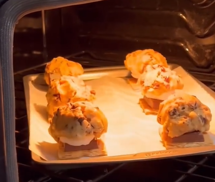 Chocolate chip cookie dough sandwiches with marshmallows and chocolate pieces are baking on a tray lined with parchment paper inside an oven.