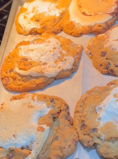 Six large chocolate chip cookies topped with melted, slightly browned marshmallows are baking on a parchment-lined tray in the oven. The cookies appear golden brown and gooey.