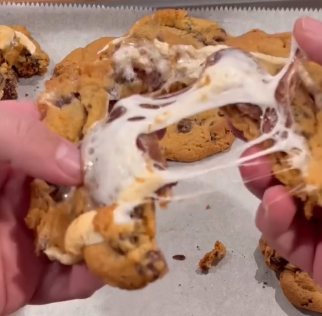 A close-up of hands pulling apart a gooey chocolate chip cookie, with melted marshmallow stretching between the halves, on a baking tray with other cookies in the background.