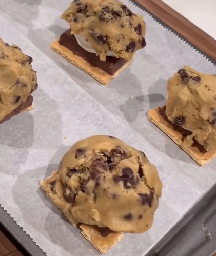 Four graham cracker squares topped with chocolate, marshmallow, and large scoops of chocolate chip cookie dough are arranged on a parchment-lined baking sheet, ready to be baked.