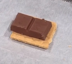 A piece of chocolate with a bite taken out of it sits on top of a square graham cracker, placed on a sheet of parchment paper.