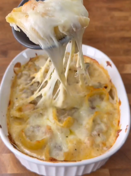 A cheesy baked casserole in a white dish, with a serving spoon lifting out a portion, showing stretchy melted cheese. The casserole sits on a wooden surface.