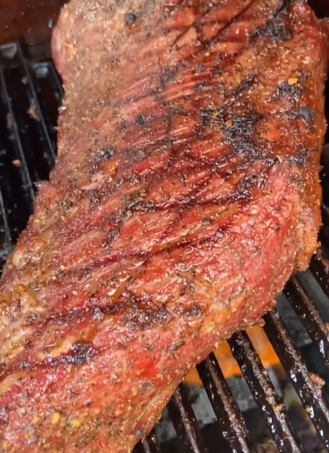 A large, seasoned steak with visible grill marks is cooking on a barbecue grill over an open flame. The meat has a browned, crispy exterior and appears juicy and well-cooked.