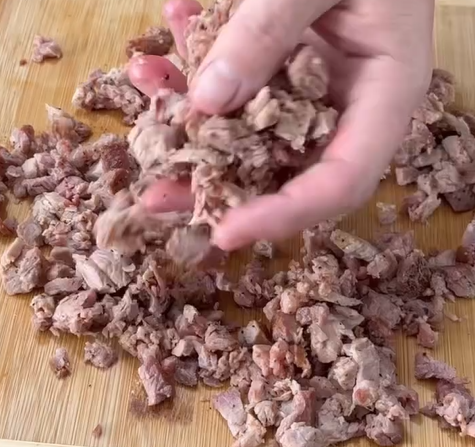 A hand holds a portion of chopped cooked meat over a wooden cutting board, with more pieces of meat scattered on the board below.