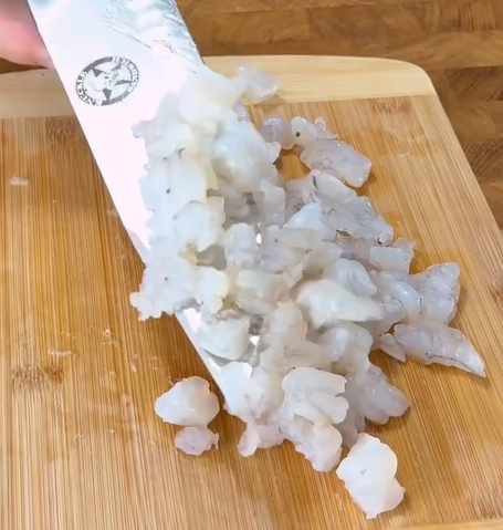 A close-up of a knife lifting a pile of diced raw fish from a wooden cutting board. The fish appears translucent and white, and the scene suggests food preparation.