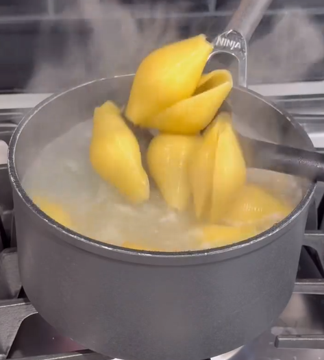 Large pasta shells boiling in a pot of hot water on a stove, with steam rising and a utensil stirring the pasta.