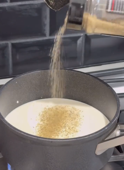 A hand sprinkles ground black pepper from a jar into a pot of creamy white liquid on a stovetop, with a black tile backsplash in the background.