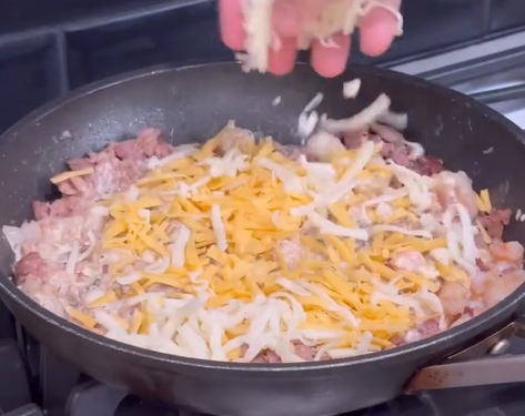 A hand sprinkles shredded cheese onto a pan filled with browned ground meat, cooking on a stove.