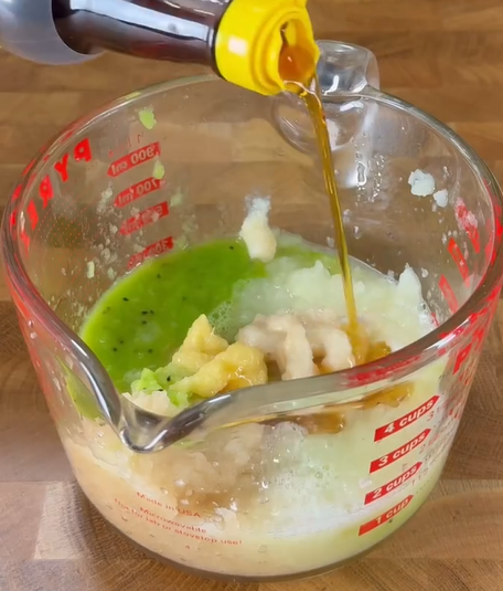 A measuring cup on a wooden surface contains blended green and beige mixtures, likely fruit or vegetable purees. Soy sauce is being poured from a bottle with a yellow cap into the cup.