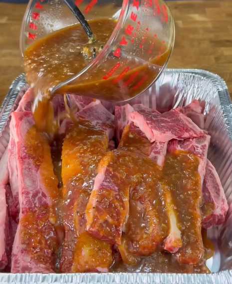 A measuring cup pours a thick, brown marinade over raw beef short ribs arranged in a foil tray on a wooden surface.