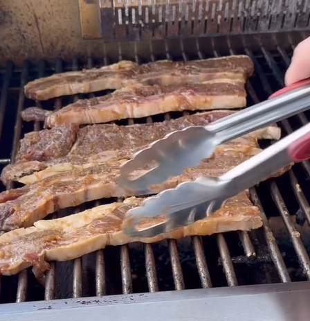 Four strips of marinated meat are being grilled on a barbecue. A hand with red-painted nails is holding metal tongs, about to turn or move one of the pieces of meat.