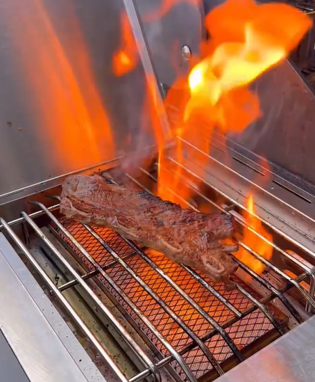 A piece of steak is being grilled over open flames on a stainless steel barbecue grill, with bright orange fire rising around the meat.