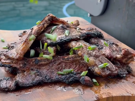 A pile of grilled beef short ribs garnished with chopped green onions sits on a wooden cutting board, with a grill and a pool in the background.