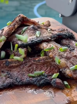 Grilled beef short ribs garnished with chopped green onions are stacked on a wooden cutting board, with a blurred background showing a pool and grill.