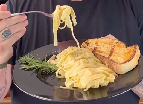 A person holds a black plate with fettuccine pasta, a slice of grilled bread, and a sprig of rosemary. The person is lifting some pasta with a fork and is wearing a black shirt and a ring.