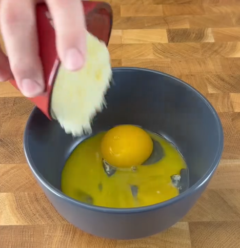 A hand pours shredded cheese from a small red cup into a dark bowl containing a cracked raw egg on a wooden surface.