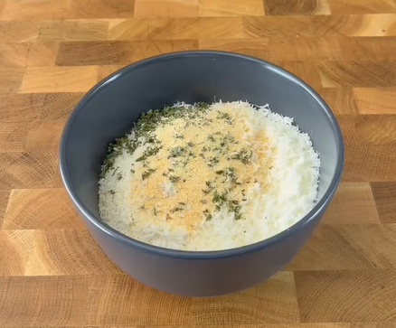 A dark gray bowl filled with finely grated cheese, garnished with dried herbs and seasoning, sits on a wooden surface.