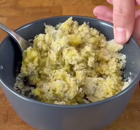 A hand holding a gray bowl filled with a mixture of mashed bananas. A spoon is resting inside the bowl, and the bowl is placed on a wooden surface.