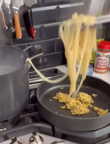 Tongs lift cooked pasta from a pot of boiling water into a pan with a clump of pasta sauce, on a stove. Knives hang on the wall, and a can of seasoning sits nearby.