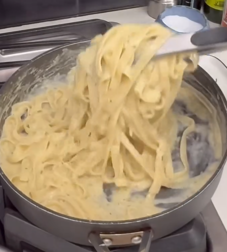 Fettuccine pasta is being stirred in a large pan with creamy Alfredo sauce on a stovetop. The creamy sauce coats the noodles as they are lifted with kitchen tongs.