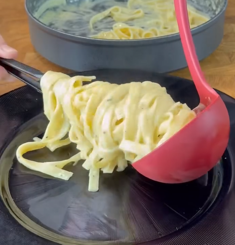 A serving of creamy fettuccine pasta is being placed onto a black plate using a red ladle, with more pasta visible in a pan in the background.