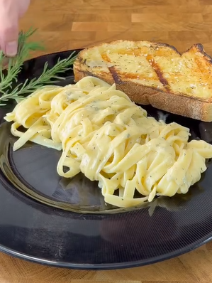 A black plate holds creamy fettuccine pasta and a slice of grilled garlic bread. A hand reaches in from the side, placing a sprig of fresh rosemary on the plate. The background is a wooden surface.
