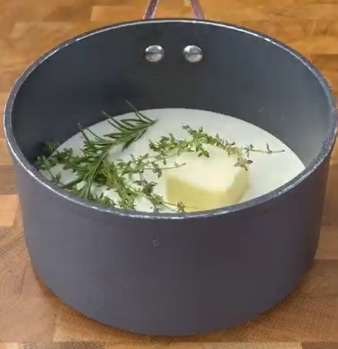 A black saucepan containing milk, a large pat of butter, and sprigs of fresh thyme and rosemary sits on a wooden countertop.
