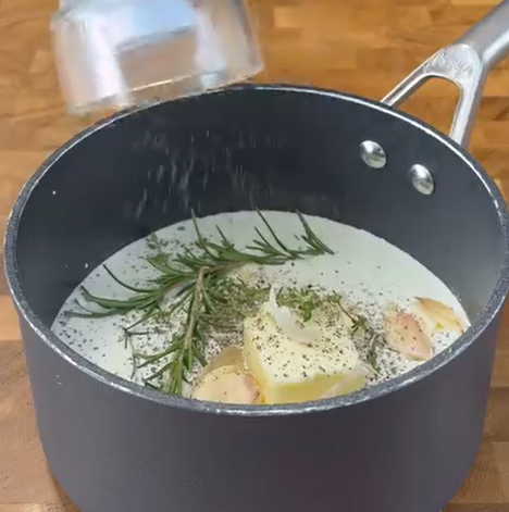 A saucepan contains cream, sprigs of rosemary, garlic, lemon wedges, and ground black pepper, with additional seasoning being sprinkled in from above. The pot sits on a wooden surface.
