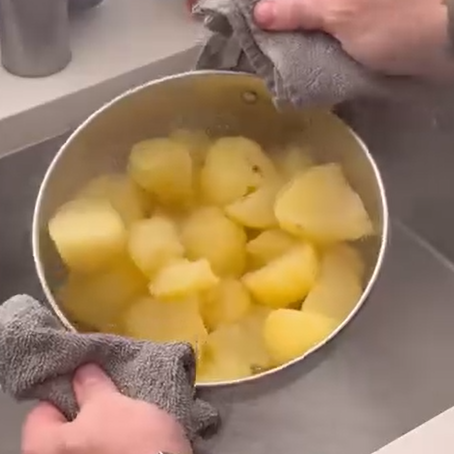 Hands holding a pot with steaming, peeled, boiled potatoes over a sink, using grey towels to grip the hot pot.