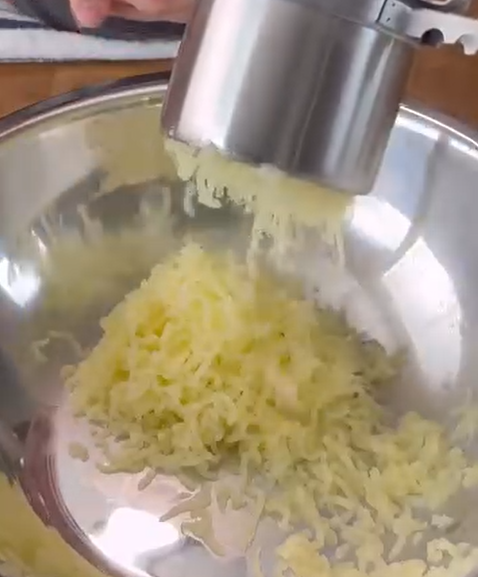 A close-up of grated potatoes being pressed into a large metal mixing bowl, likely using a potato ricer or grater.