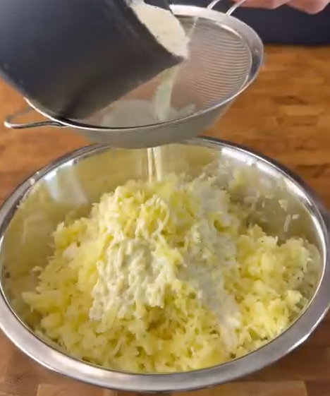 Shredded potatoes in a metal bowl with flour being sifted over them from a strainer, on a wooden surface.