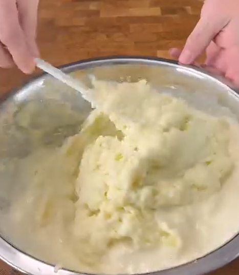 A person is mixing mashed potatoes in a large metal bowl with a spoon, on a wooden surface.