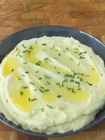 A bowl of creamy mashed potatoes topped with melted butter and chopped chives, placed on a wooden surface.