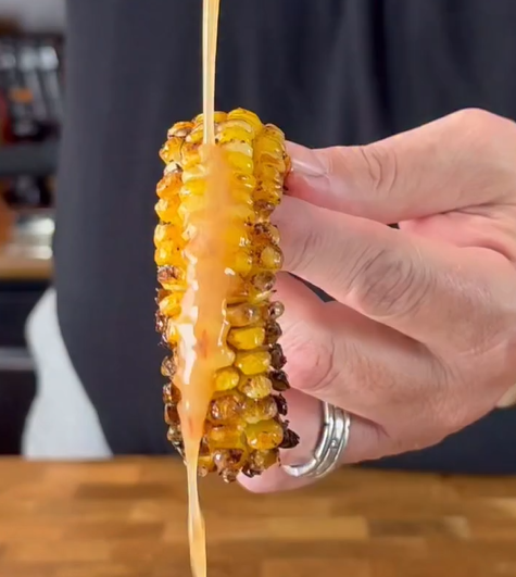 A hand holds a grilled corn on the cob while a stream of sauce is poured over it, with a kitchen background visible.
