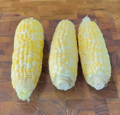 Three ears of partially shucked corn with pale yellow and white kernels are placed side by side on a wooden cutting board.