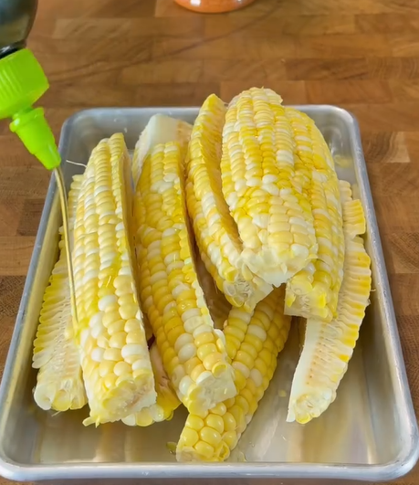 A tray of raw corn on the cob cut into halves is arranged on a metal tray, with a green squeeze bottle pouring a liquid over the corn. The tray sits on a wooden surface.
